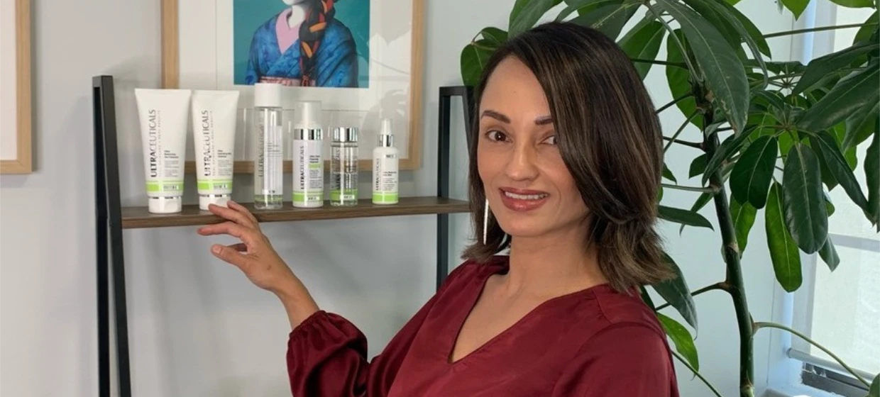 A woman in a burgundy blouse stands beside a shelf with ultraceuticals products, reaching towards them. Behind her is a large leafy plant and a framed colorful portrait on the wall.